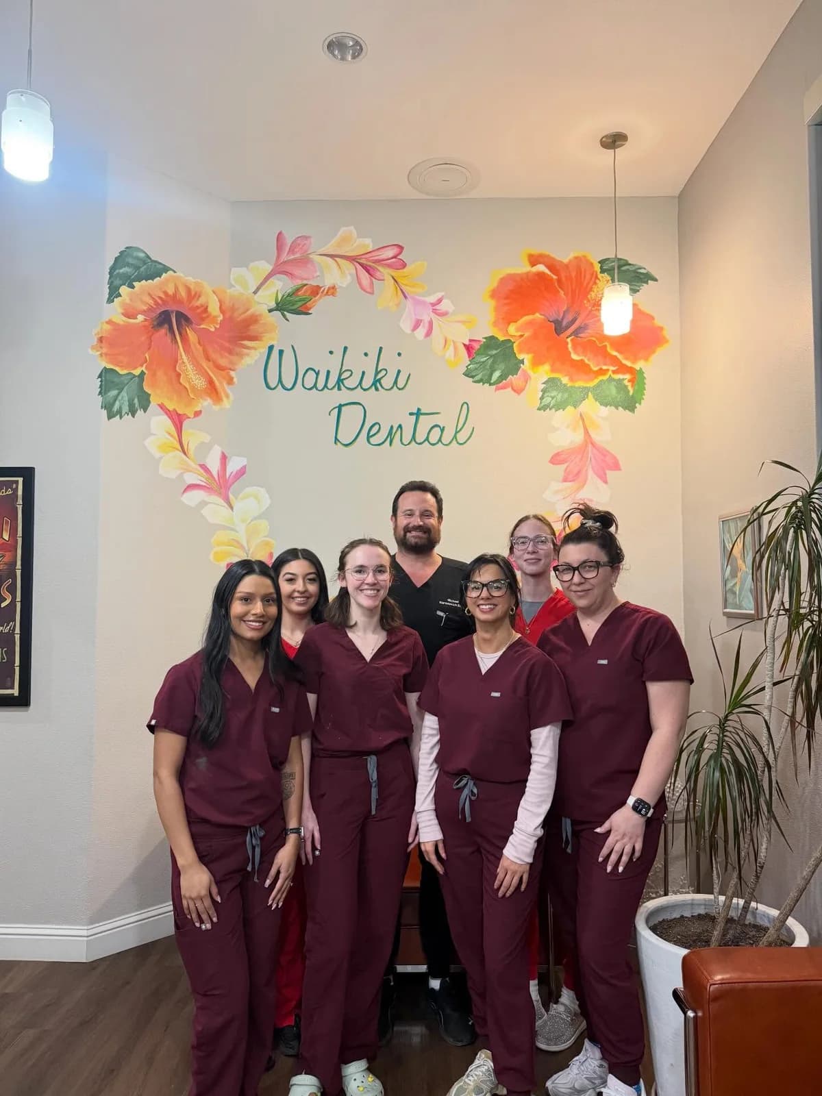 Dr. Michael Narodovich with the Waikiki Dental team in burgundy scrubs in front of the practice's hibiscus mural